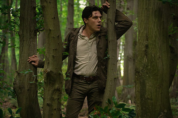 A man in a brown jacket peeks cautiously from behind thick tree trunks in a dense green forest.