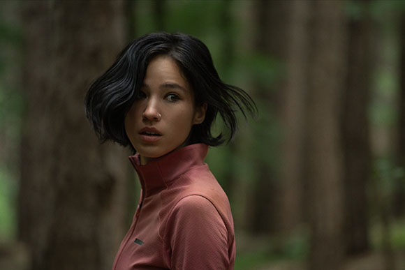 A young woman with short dark hair looks back with a concerned expression in a wooded area.