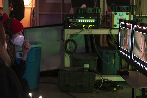A person wearing a red beanie and white face mask watches video monitors in a dark production studio.