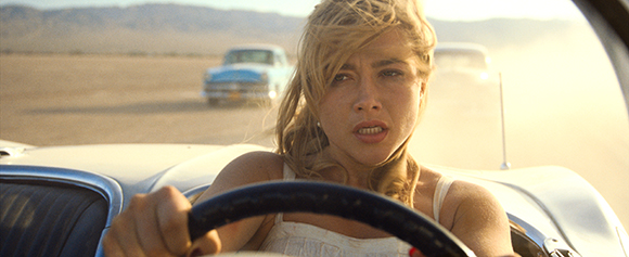 A blonde woman with a concerned expression drives a white convertible through a dusty desert.