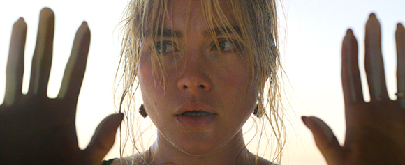 A blonde woman with wet hair looks through a glass surface with both hands pressed against it.