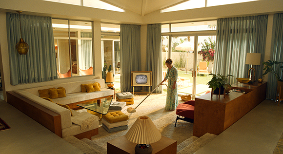 A person in a patterned dress vacuums a light-colored rug in a sunlit, mid-century modern living room.