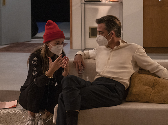 A person in a red beanie and a person in a white shirt sit on a couch wearing face masks.