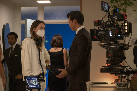 A woman in a white hoodie and mask stands near a man in a suit and a professional film camera.