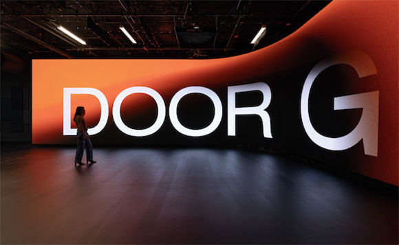 A person stands in a dark room looking up at a large, curved orange and black screen with the text 'DOOR G'.