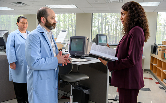 A woman in a maroon suit holds a document while talking to two people in light blue lab coats.