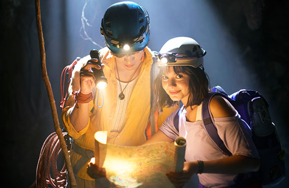 Two people wearing helmets with headlamps look at a map in a dark cave.