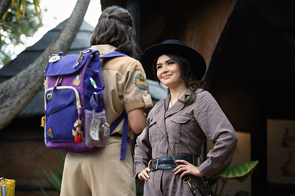A smiling woman in a brown jumpsuit and black hat stands beside a person in a tan uniform with a purple backpack.