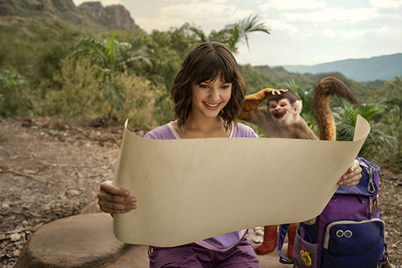 A smiling young woman holds a large parchment scroll while a small monkey sits beside her in a tropical landscape.