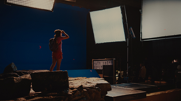 A person in a red shirt and shorts stands before a large blue screen on a film set with bright studio lights.