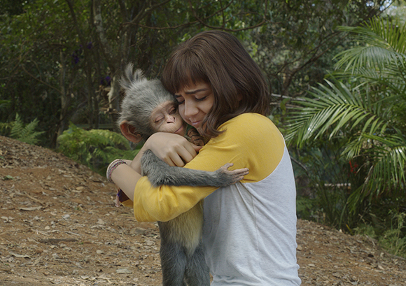 A young woman with brown hair wearing a yellow and white shirt hugs a small grey monkey.