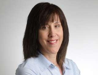 A woman with shoulder-length dark hair and bangs wearing a light blue striped shirt, smiling against a white background.