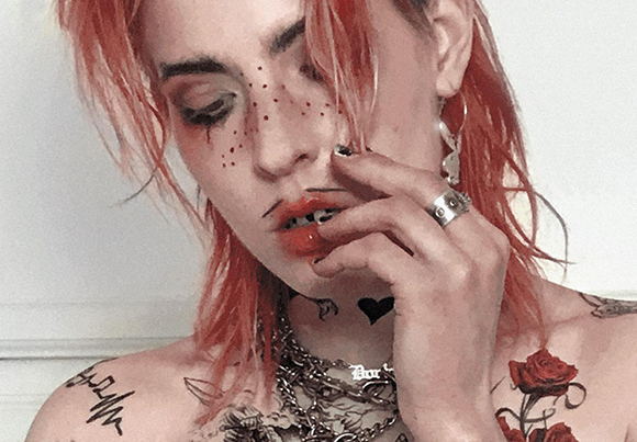 A person with pink hair, facial freckles, and various tattoos looking down with a hand near their mouth.