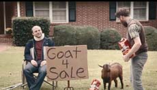 A man sits by a sign reading 'Goat 4 Sale' as another man approaches a small brown goat in a yard.