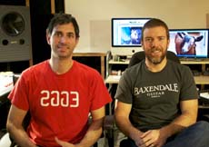 Two men sit side by side in a room with computer monitors in the background.