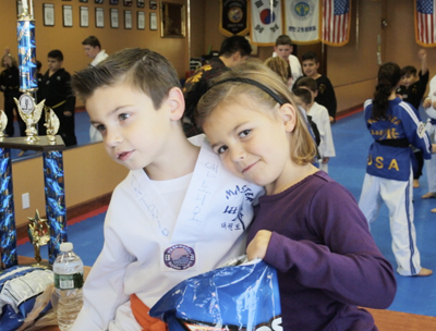 A young boy in a white martial arts uniform and a young girl in a purple shirt hug closely.