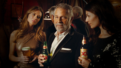 A man in a suit holding a beer bottle stands between two women holding drinks in a dark lounge.