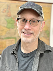 A man with glasses and a grey baseball cap smiling in front of a map.
