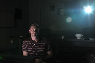 A man in a striped polo shirt sits in a dark room lit by a single bright light from the upper right.