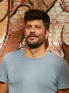 A man with dark hair and a beard wearing a light blue t-shirt, smiling in front of a graffiti brick wall.