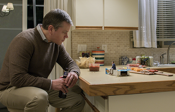 A man in a brown sweater sits at a kitchen counter, staring intently at a miniature person standing near food.