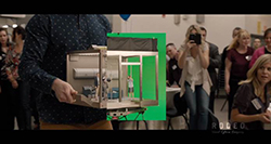 A person holds a small scale model of a room in front of a green screen in a crowded room.