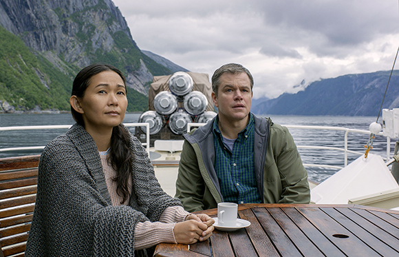 A woman in a grey shawl and a man in a green jacket sit at a wooden table on a boat near mountains.