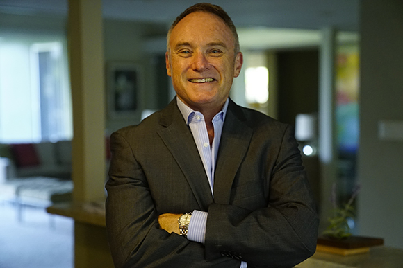 A smiling man in a dark suit and light blue shirt stands with his arms crossed in an indoor setting.
