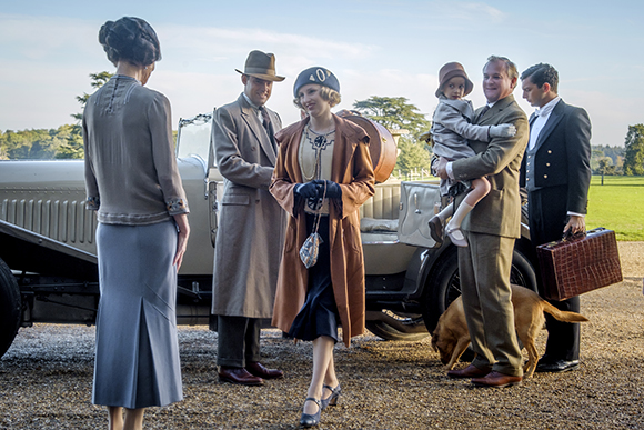 People in early 20th-century clothing stand near a vintage car on a gravel path.