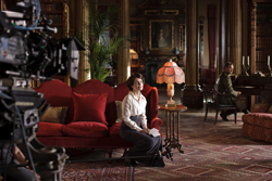 A film crew shoots a scene of a woman on a red sofa and a man sitting at a desk in a classic library.
