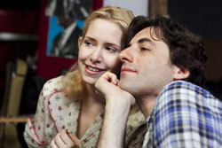 A blonde woman in a floral shirt leans her head against a dark-haired man in a plaid shirt.