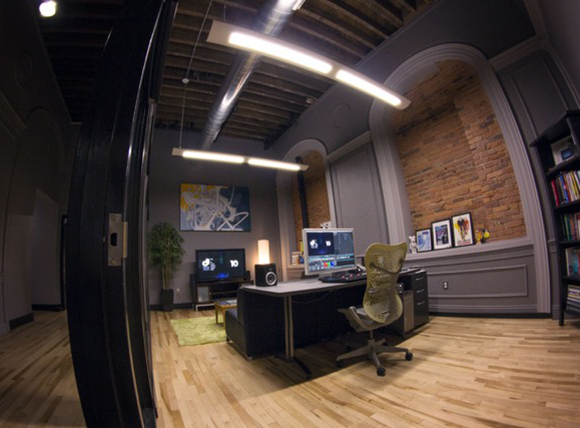 A home office featuring a large desk, computer monitors, an ergonomic chair, and exposed brick walls.
