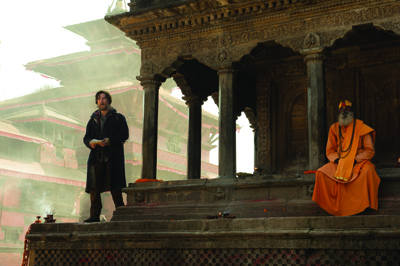 A man in dark clothing stands on a stone ledge near a monk in orange robes by an ancient temple.