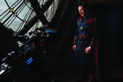 A man in a blue tunic and long red cloak stands in a dark room next to a large window and mechanical equipment.