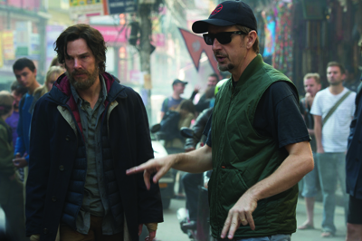 A man in a black jacket walks beside a man in a green vest and sunglasses in a busy street.