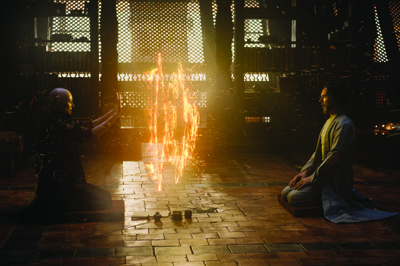 Two men sit facing each other in a dark room with a vertical column of fire between them.