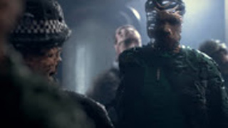 A person wearing dark, textured armor stands in a dark, smoky room near other figures.