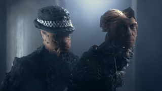 Two uncanny, disfigured humanoid figures wearing dark clothing in a dimly lit, smoky room.