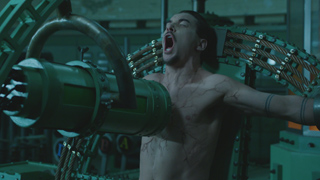 A shirtless man with his mouth wide open in a scream, strapped into a large green mechanical machine.