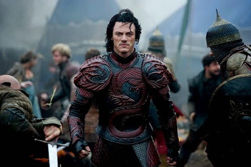 A man in ornate red and black dragon-scale armor stands in a crowded, smoky battlefield setting.