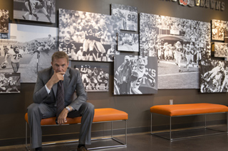 A man in a grey suit sits on an orange bench before a wall covered in large black and white sports photos.