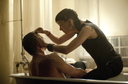A woman in a black tank top combs the hair of a shirtless man sitting in a bathtub.