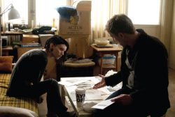 A man and woman sit in a room filled with boxes and papers, looking down at documents.