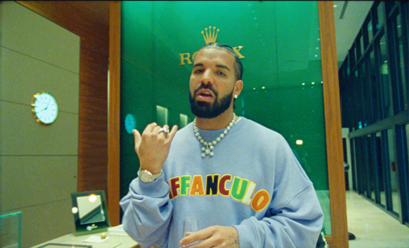 A man with a beard wearing a light blue sweatshirt and diamond necklaces stands before a green wall.