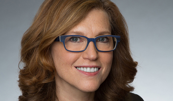A woman with wavy reddish-brown hair and blue-rimmed glasses smiling at the camera.