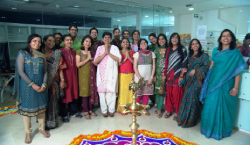A group of women in colorful sarees and kurtas posing around a lit brass lamp and rangoli.