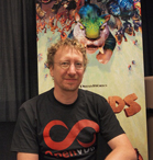 Man with curly hair and glasses wearing a black t-shirt with a red infinity symbol smiles in front of a movie poster.