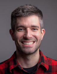 A smiling man with short brown hair and stubble wearing a red and black checkered shirt.
