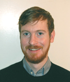 A man with reddish hair and a beard smiling against a plain white background.