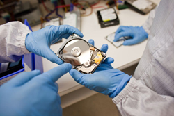 Two people in blue gloves and white lab coats examine a small hard drive component on a workbench.
